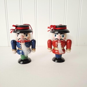 May include: Two wooden nutcrackers, each with a black hat, red accents, and a white beard. One wears a blue uniform, the other a red uniform. They stand on a white surface against a white paneled wall.