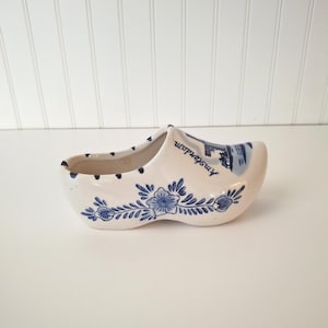 May include: A ceramic Dutch clog in white with blue floral and Amsterdam imagery. The decorative shoe features a blue and white windmill and canal scene. The clog is a traditional Dutch souvenir.