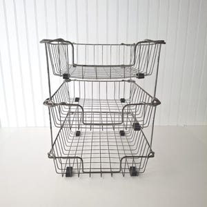 May include: A three-tiered, wire-frame storage basket. The metal baskets are rectangular with rounded corners and a grid pattern. The frame is a dark gray color, and the baskets are stacked on top of each other. The background is white.