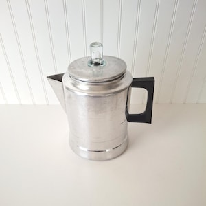 May include: A vintage aluminum coffee percolator with a black handle and a glass knob on the lid. The coffee pot has a spout and is sitting on a white surface. The background is a white wall with vertical paneling.