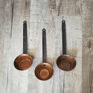 May include: Three matching measuring ladles featuring round, polished copper bowls and long, flat, dark iron handles. Each handle includes a circular hole at the top for hanging. The copper bowls have a warm, metallic finish, while the iron handles appear dark, matte, and sturdy, providing a clear contrast in texture and color.