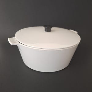 May include: A white, oval-shaped casserole dish with a matching lid and a black knob. The dish has two small handles on either side. The lid fits snugly on top of the dish. The casserole dish is made of ceramic or porcelain.