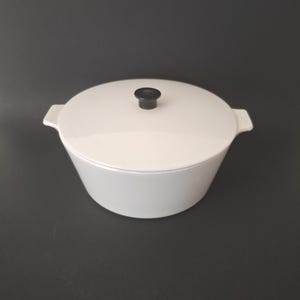 May include: A white, lidded casserole dish with a black knob. The dish has a rounded shape with two side handles. The lid sits flush on the dish, and the knob is centered on the lid. The casserole dish is on a dark gray background.