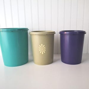 May include: Three vintage plastic containers in teal, tan, and purple. The teal container is slightly angled. The tan container has a white floral design. All containers have lids and a cylindrical shape, perfect for storage.
