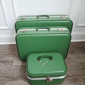 May include: A set of three green suitcases of varying sizes, each featuring a green handle, silver-toned metal latches, and white trim. The smallest suitcase is positioned in the front and includes a small silver key resting on its top surface.