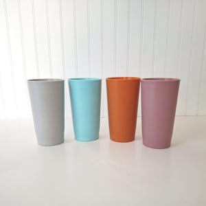 May include: Four tall, cylindrical tumblers in a row. The tumblers are in shades of gray, turquoise, orange, and mauve. The tumblers are made of a solid material and have a smooth, matte finish. The background is a white wall.