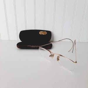 May include: Vintage eyeglasses with a gold-tone frame and clear lenses. The glasses are positioned next to an open, black eyeglass case with a gold-colored emblem. The case has a maroon interior.