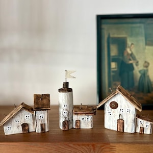 Può includere: Un set di cinque case in miniatura in legno sbiancato con tetti e dettagli marroni. Le case sono disposte in fila su una superficie in legno. Una casa ha una piccola bandiera bianca in cima.