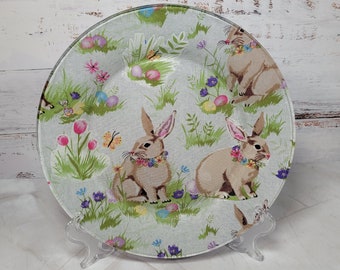 May include: A light blue glass charger plate with a floral pattern featuring bunnies, butterflies, and Easter eggs. The plate is on a clear acrylic stand.