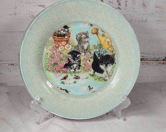 May include: A light blue glass plate with a white rim and a floral design featuring kittens and butterflies. The plate is on a white stand.