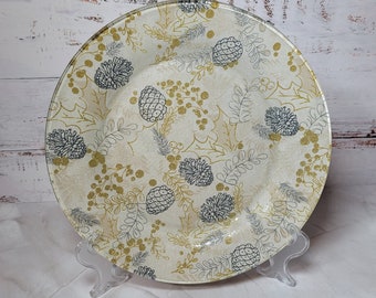 May include: A white ceramic plate with a gold and gray holiday design. The plate features a repeating pattern of pine cones, holly leaves, and berries.