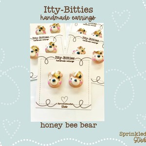 Puede incluir: Par de pendientes hechos a mano de arcilla polimérica con un diseño de oso abeja. El oso es marrón con mejillas rosadas y una abeja amarilla en la cabeza. Los pendientes están en una tarjeta blanca con el texto "Itty-Bitties handmade earrings" y "Sprinkledoodle Studio".