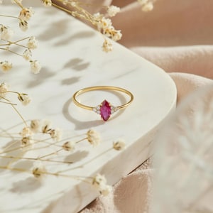 May include: A thin gold ring featuring a central marquise-cut deep pink gemstone flanked by two small clear round stones on each side, set in a delicate metal band that rests on a smooth, light-colored surface with white flowers.