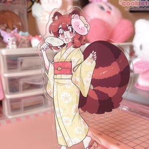 May include: A clear acrylic standee featuring a cartoon character in a yellow kimono with floral patterns, holding a fan. The character has red hair, a red and brown tail, and a pink mask. The base is blue with a fireworks design. The text "cookietanuki" is visible.