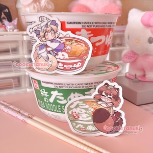 May include: Two instant ramen bowls with character stickers and a pair of chopsticks. The bowls are white with red and green text. The stickers feature anime-style characters and ramen bowl illustrations. The chopsticks are light brown with a pink pattern.