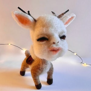 May include: A whimsical felted deer sculpture with a human-like face, featuring a white head, pink lips, and dark eyes. The deer has small, dark antlers and a brown and white body. The sculpture is set against a white background.