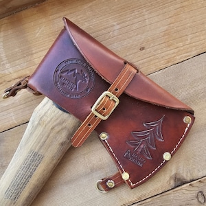 May include: A brown leather axe sheath with a belt strap and a brass buckle. The sheath has a stamped design of a pine tree and the text "Idaho Made". The sheath is attached to a wooden axe handle with a black axe head.