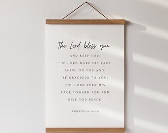 May include: A minimalist wall art print featuring the text "The Lord bless you". The white print with black text is held by a wooden frame and a brown string. The artwork is displayed against a white wall, with the text "Numbers 6:24-26".