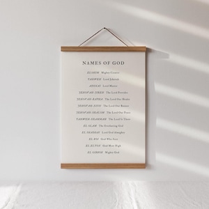 May include: A framed print with the words "NAMES OF GOD" at the top, followed by a list of names and their meanings. The print is cream-colored with black text and wooden bars at the top and bottom, suspended by a brown cord.