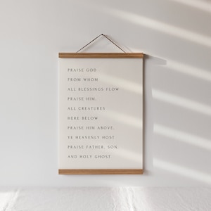 May include: A framed canvas print with the text "PRAISE GOD FROM WHOM ALL BLESSINGS FLOW PRAISE HIM, ALL CREATURES HERE BELOW PRAISE HIM ABOVE. YE HEAVENLY HOST PRAISE FATHER, SON, AND HOLY GHOST." The frame is made of light brown wood.
