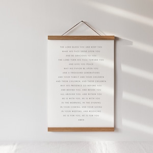 May include: A white canvas print with black text, held by a wooden frame and a hanging cord. The text is a religious blessing. The print is displayed against a white wall, with shadows.
