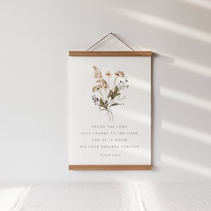 May include: A framed print with a floral arrangement and the text "PRAISE THE LORD. GIVE THANKS TO THE LORD. FOR HE IS GOOD: HIS LOVE ENDURES FOREVER. PSALM 106:1." The frame is made of light-colored wood, and the print is hanging on a white wall.