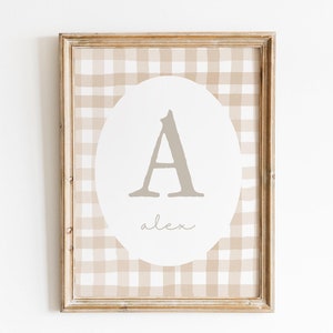 May include: Framed wall art featuring a tan and white gingham pattern background. A white oval in the center displays the letter "A" in a muted gray color, with the name "alex" written below in script. The frame is a light brown.