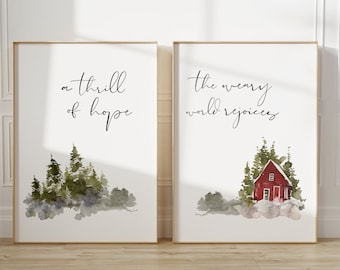 May include: Two framed art prints with gold frames. One print reads "a thrill of hope" above a watercolor image of evergreen trees. The other print reads "the weary world rejoices" above a red cabin.