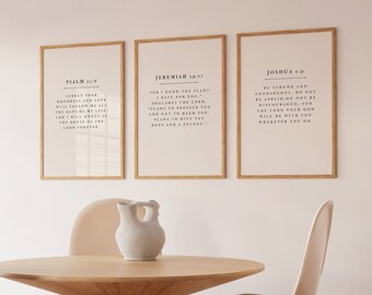 May include: Three framed prints with Bible verses are displayed above a wooden table and chairs. The prints feature black text on a light background, within thin wooden frames. A white vase is on the table. The room has a neutral colour palette.