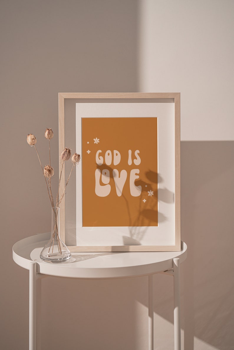 May include: A framed print with the words "GOD IS LOVE" in white, set against an orange background. The print is in a light wood frame and rests on a white table. A clear glass vase with dried flowers is on the table.