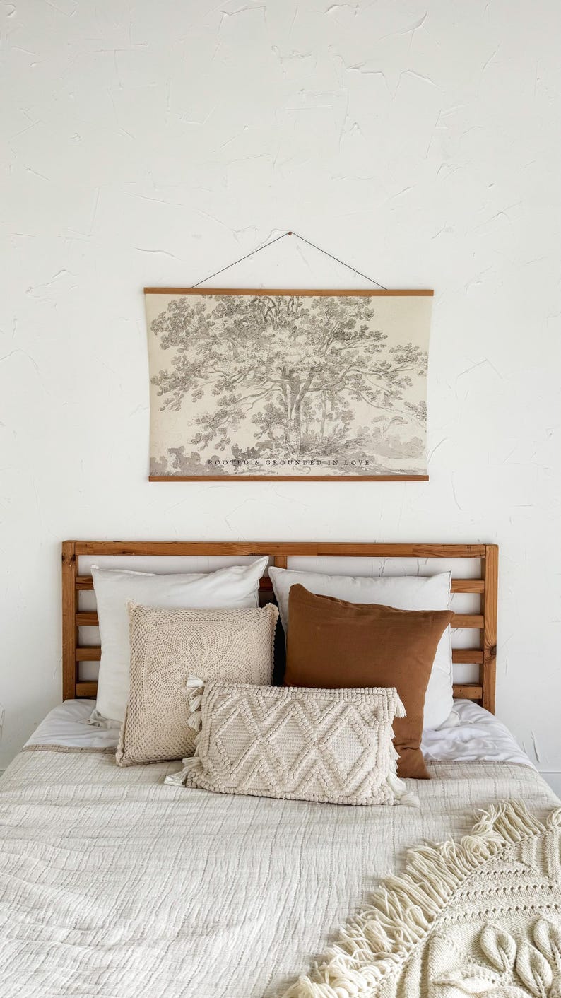 Puede incluir: Un dormitorio con cabecero de madera y una cama cubierta con un edred&oacute;n blanco. Cojines decorativos en tonos crema y marr&oacute;n est&aacute;n colocados sobre la cama. Una impresi&oacute;n enmarcada de un &aacute;rbol, con el texto "ROOTED & GROUNDED IN LOVE", cuelga sobre la cama.