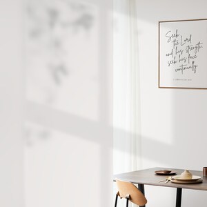 May include: A framed print with a white background and black text that reads "Seek the Lord and his strength, and seek his face continually. 1 Chronicles 16:11". The print is hanging on a white wall above a wooden table with two chairs and a vase with branches.