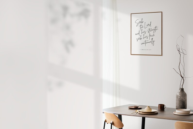 May include: A framed print with a white background and black text that reads "Seek the Lord and his strength, and seek his face continually. 1 Chronicles 16:11". The print is hanging on a white wall above a wooden table with two chairs and a vase with branches.