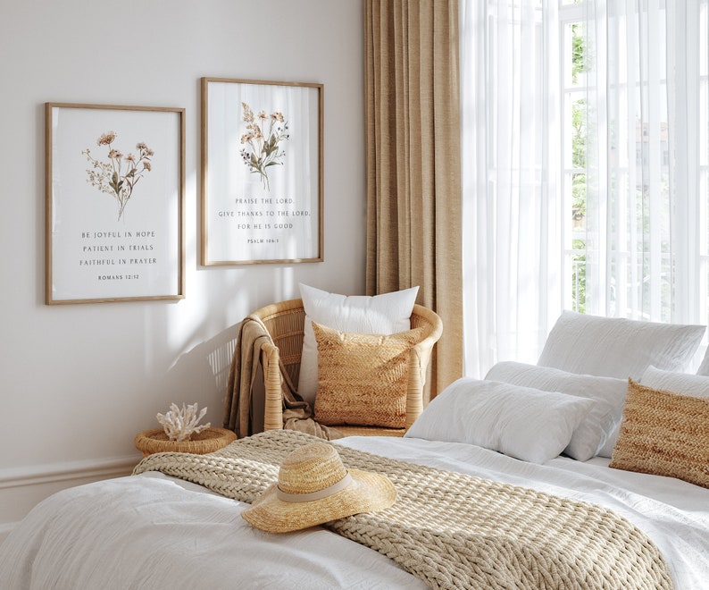 May include: Two framed botanical prints with inspirational quotes hang above a wicker chair and bed. The prints feature dried flowers and the text "BE JOYFUL IN HOPE" and "PRAISE THE LORD". A straw hat and decorative pillows add to the cozy bedroom decor.