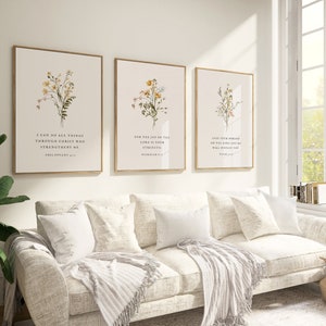 May include: Three framed botanical art prints with gold frames hang above a white sofa. Each print features a floral illustration and a Bible verse. The room is bright with natural light from a large window.