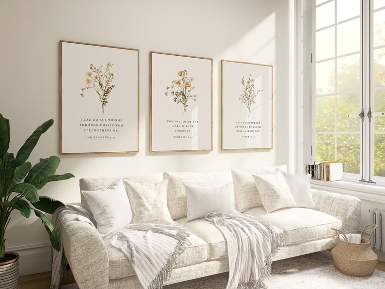May include: Three framed botanical art prints with gold frames hang above a white sofa. Each print features a floral illustration and a Bible verse. The room is bright with natural light from a large window.