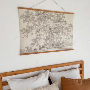 Puede incluir: Un estampado en blanco y negro de un &aacute;rbol colgado en la pared, con varillas de madera en la parte superior e inferior. El estampado est&aacute; sobre una estructura de cama de madera con almohadas blancas y marrones. El texto "ROOTED & GROUNDED IN LOVE".