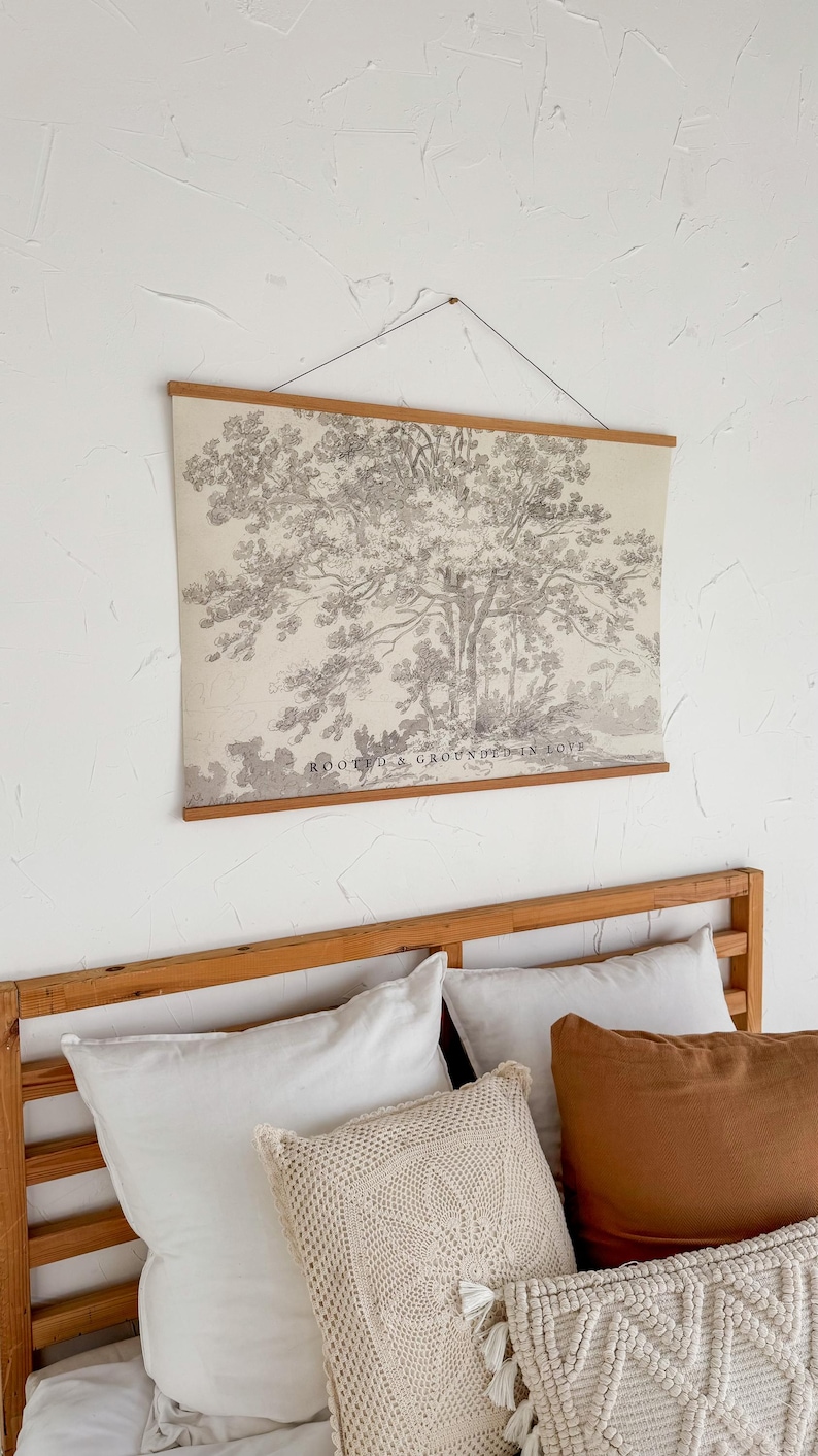 Puede incluir: Un estampado en blanco y negro de un &aacute;rbol colgado en la pared, con varillas de madera en la parte superior e inferior. El estampado est&aacute; sobre una estructura de cama de madera con almohadas blancas y marrones. El texto "ROOTED & GROUNDED IN LOVE".