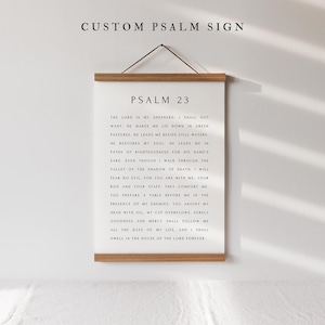 May include: A framed print with the text "CUSTOM PSALM SIGN" at the top and "PSALM 23" below. The text of Psalm 23 is printed in black on a white background. The frame is made of light-colored wood, and the print hangs from a brown leather cord.