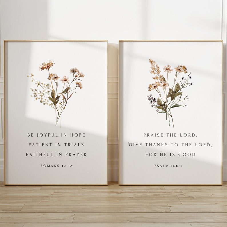 May include: Two framed art prints with floral watercolor designs. The left print features text from Romans 12:12, and the right print features text from Psalm 106:1. Both prints have gold frames and are displayed on a white background.