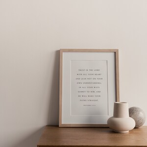 May include: A framed print with a white background and black text. The text reads "Trust in the Lord with all your heart and lean not on your own understanding; in all your ways submit to him, and he will make your paths straight. Proverbs 3:5-6". The print is sitting on a wooden surface with a small white vase and a round stone.