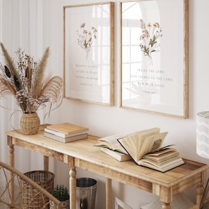 May include: Two framed art prints with botanical illustrations and inspirational quotes. The prints are displayed on a light wood console table with books and decorative items. The text on the left print reads: "BE JOYFUL IN HOPE, PATIENT IN TRIALS, FAITHFUL IN PRAYER, ROMANS 12:12."