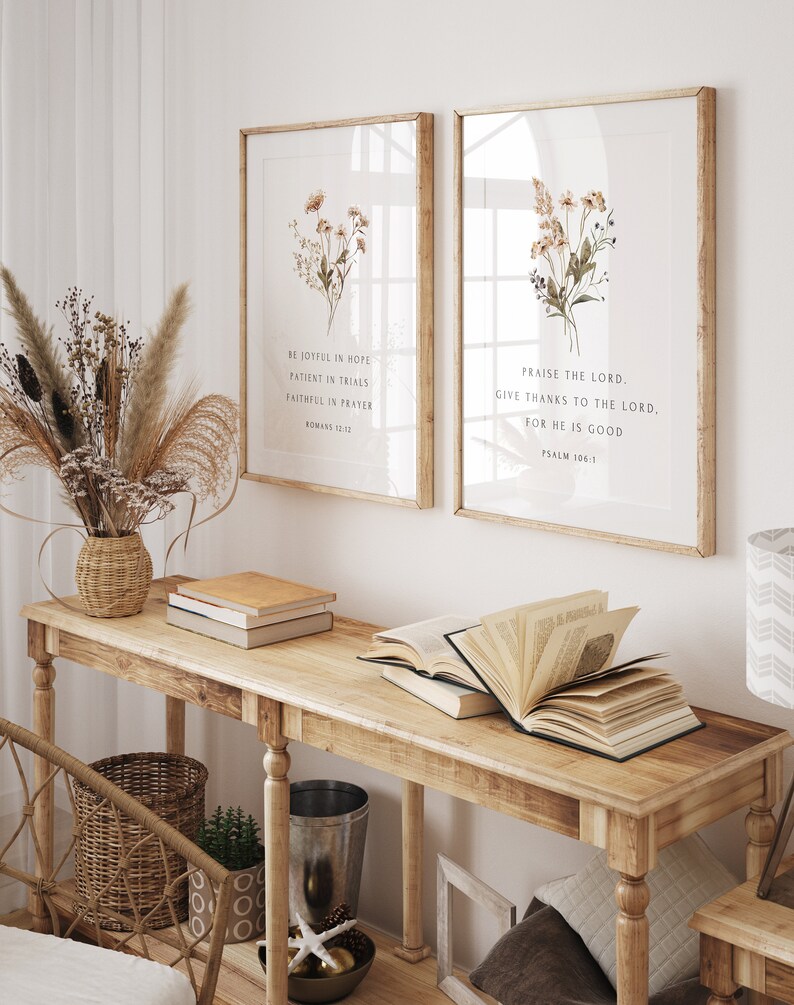 May include: Two framed art prints with botanical illustrations and inspirational quotes. The prints are displayed on a light wood console table with books and decorative items. The text on the left print reads: "BE JOYFUL IN HOPE, PATIENT IN TRIALS, FAITHFUL IN PRAYER, ROMANS 12:12."