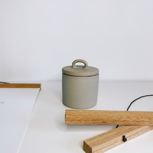 May include: A small, speckled gray ceramic jar with a lid and a handle. The jar is cylindrical and sits on a white surface. Wooden poster hangers are also visible.