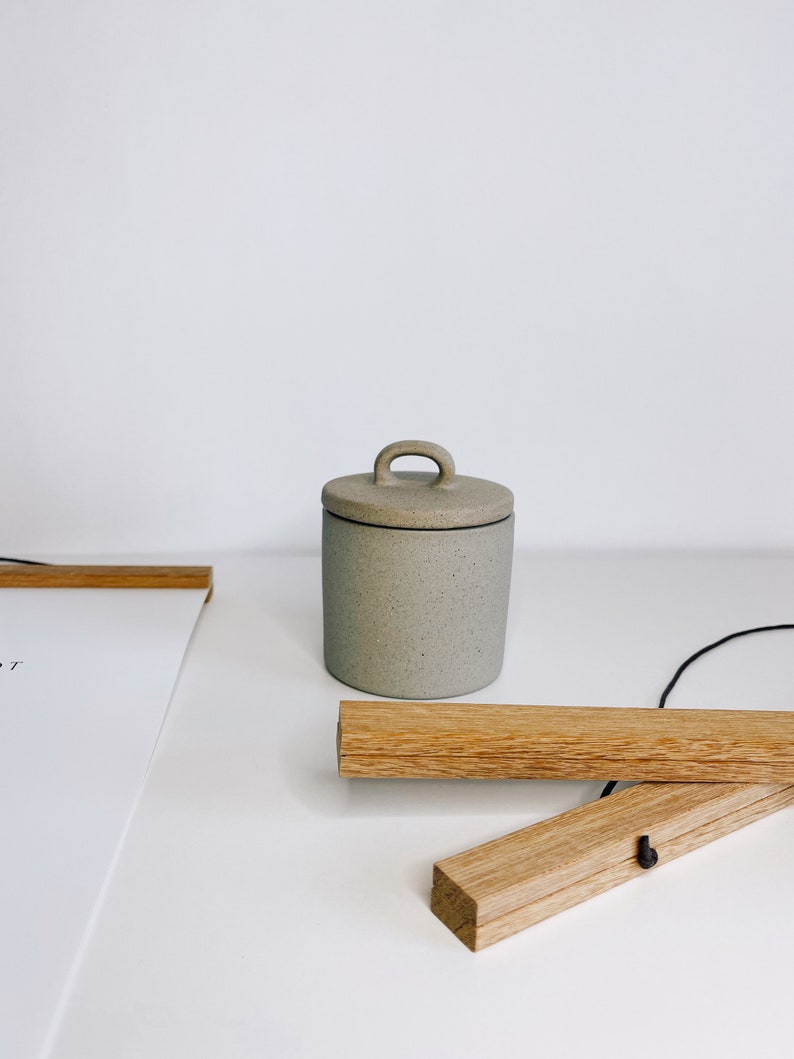 May include: A small, speckled gray ceramic jar with a lid and a handle. The jar is cylindrical and sits on a white surface. Wooden poster hangers are also visible.