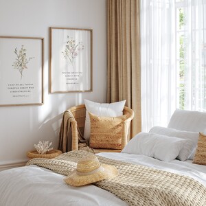 May include: Two framed botanical prints with inspirational quotes hang above a wicker chair and bed. The prints feature floral arrangements and text. A straw hat rests on the bed, alongside pillows and a woven throw. The room is bathed in natural light.