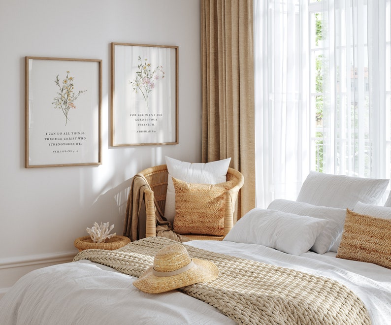 May include: Two framed botanical prints with inspirational quotes hang above a wicker chair and bed. The prints feature floral arrangements and text. A straw hat rests on the bed, alongside pillows and a woven throw. The room is bathed in natural light.