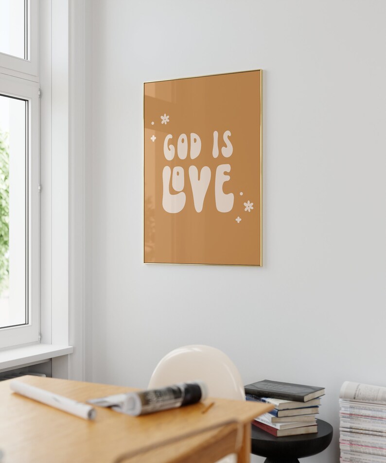 May include: A framed art print with the words "GOD IS LOVE" in white, stylized lettering on a tan background. The print is in a gold frame and is hanging on a white wall. Small white floral accents are on the print.