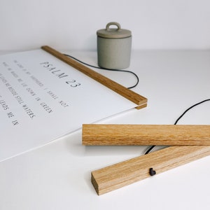 May include: A white print with the text "PSALM 23" and other scriptural text, held by two wooden magnetic poster hangers. A small, gray ceramic container with a handle sits nearby. The wooden hangers are made of light-colored wood.
