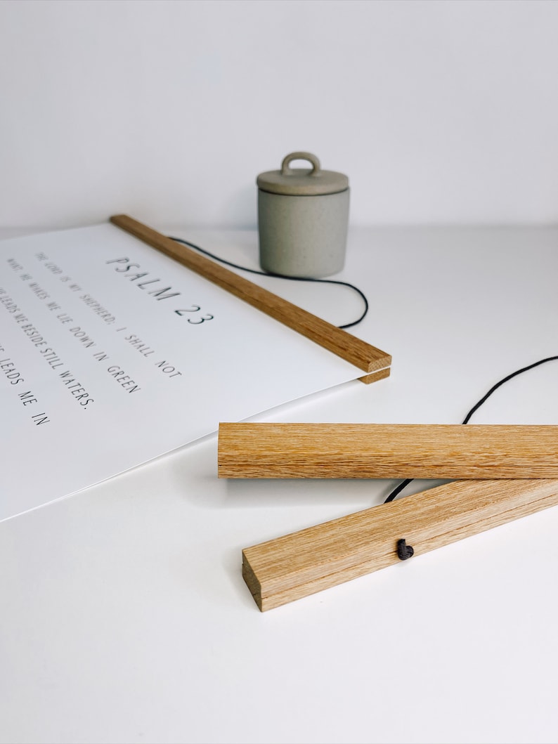 May include: A white print with the text "PSALM 23" and other scriptural text, held by two wooden magnetic poster hangers. A small, gray ceramic container with a handle sits nearby. The wooden hangers are made of light-colored wood.